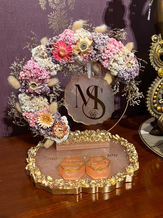 A decorative floral arrangement on a gold-toned stand with a monogram sign and two resin ring holders, displayed on a wooden surface against a purple patterned backdrop.